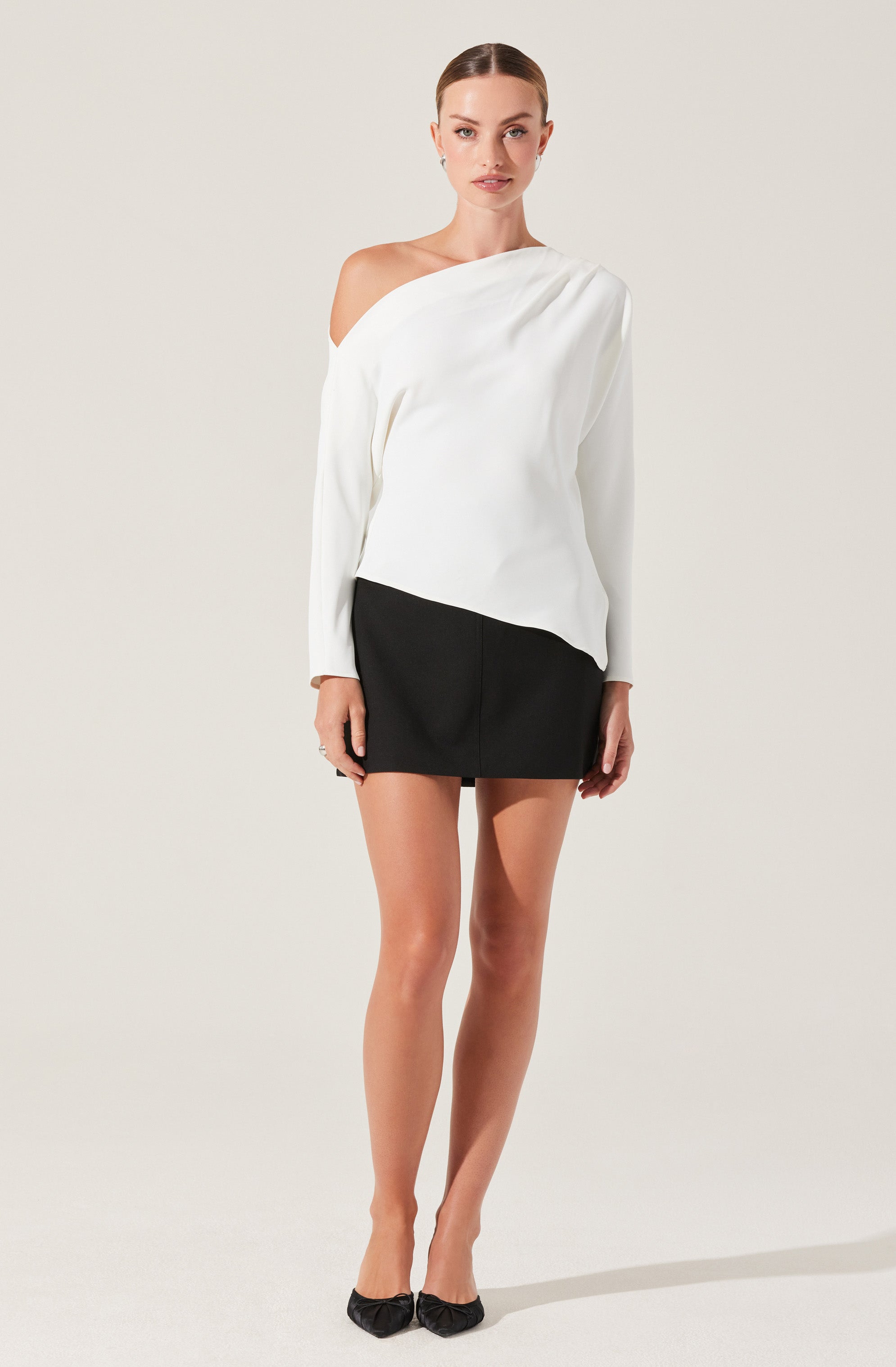A woman is wearing an off-the-shoulder white top with long sleeves, featuring a relaxed fit and pleats near the shoulder. She pairs it with a fitted, high-waisted black mini skirt. She completes the outfit with black pointed-toe shoes that have a small bow detail on top.