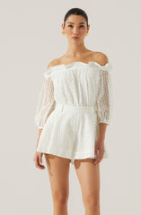 A woman is wearing an off-the-shoulder white eyelet lace top with elastic at the neckline and sleeves, and matching high-waisted shorts with a wide waistband and a flowy, relaxed fit. The top has puffed three-quarter length sleeves with a textured pattern.