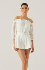 A woman is wearing an off-the-shoulder white eyelet lace outfit that consists of a loose, ruffled top with three-quarter length, puffed sleeves and high-waisted shorts. The fabric has a perforated pattern, creating a delicate, airy look. She accessorizes with large gold earrings.