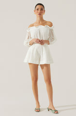 A woman wearing an off-the-shoulder white dress made of eyelet fabric with a ruffled neckline. The dress has long, semi-sheer, puffed sleeves with elastic cuffs. The matching shorts are high-waisted and also made of eyelet fabric. She is wearing light green open-toe heeled sandals and gold earrings.