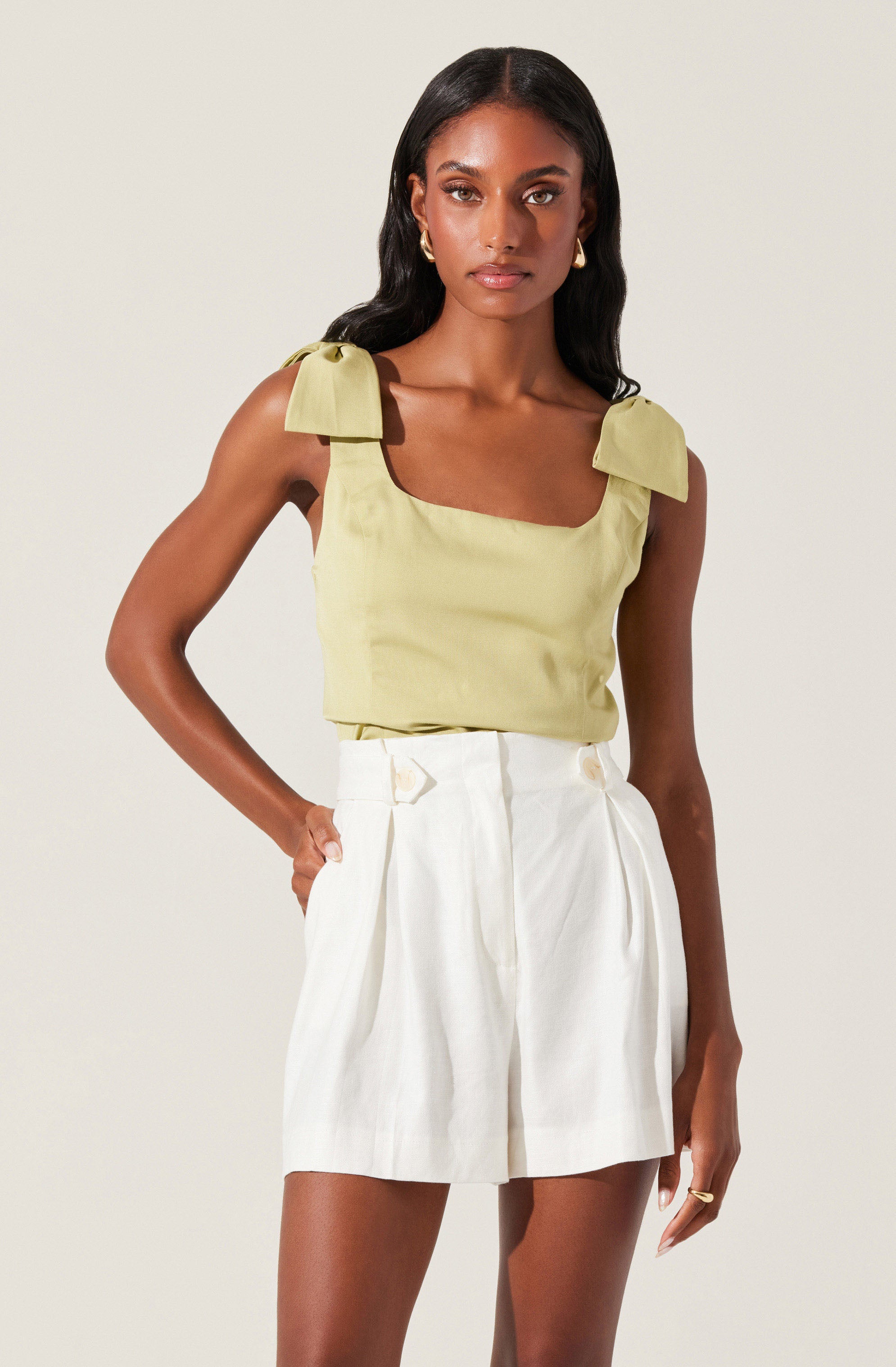 A woman is wearing a sleeveless, light yellow top with shoulder bows and wide straps. She pairs it with high-waisted, white pleated shorts that have belt loops.