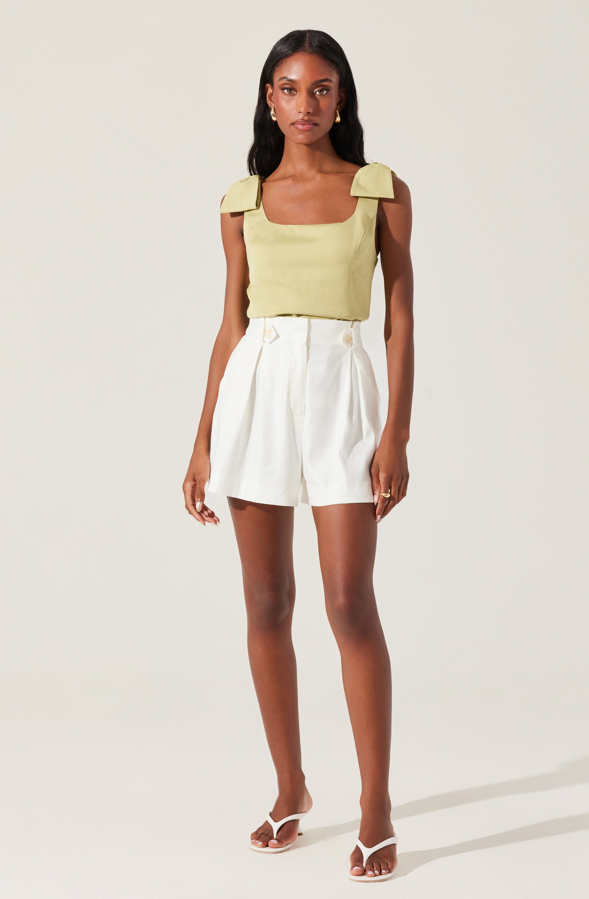 A woman is wearing a sleeveless, light yellow top with wide shoulder straps that have decorative bow details, paired with high-waisted, white tailored shorts with pleats and decorative buttons at the waist. She is also wearing white, open-toed high-heeled sandals.