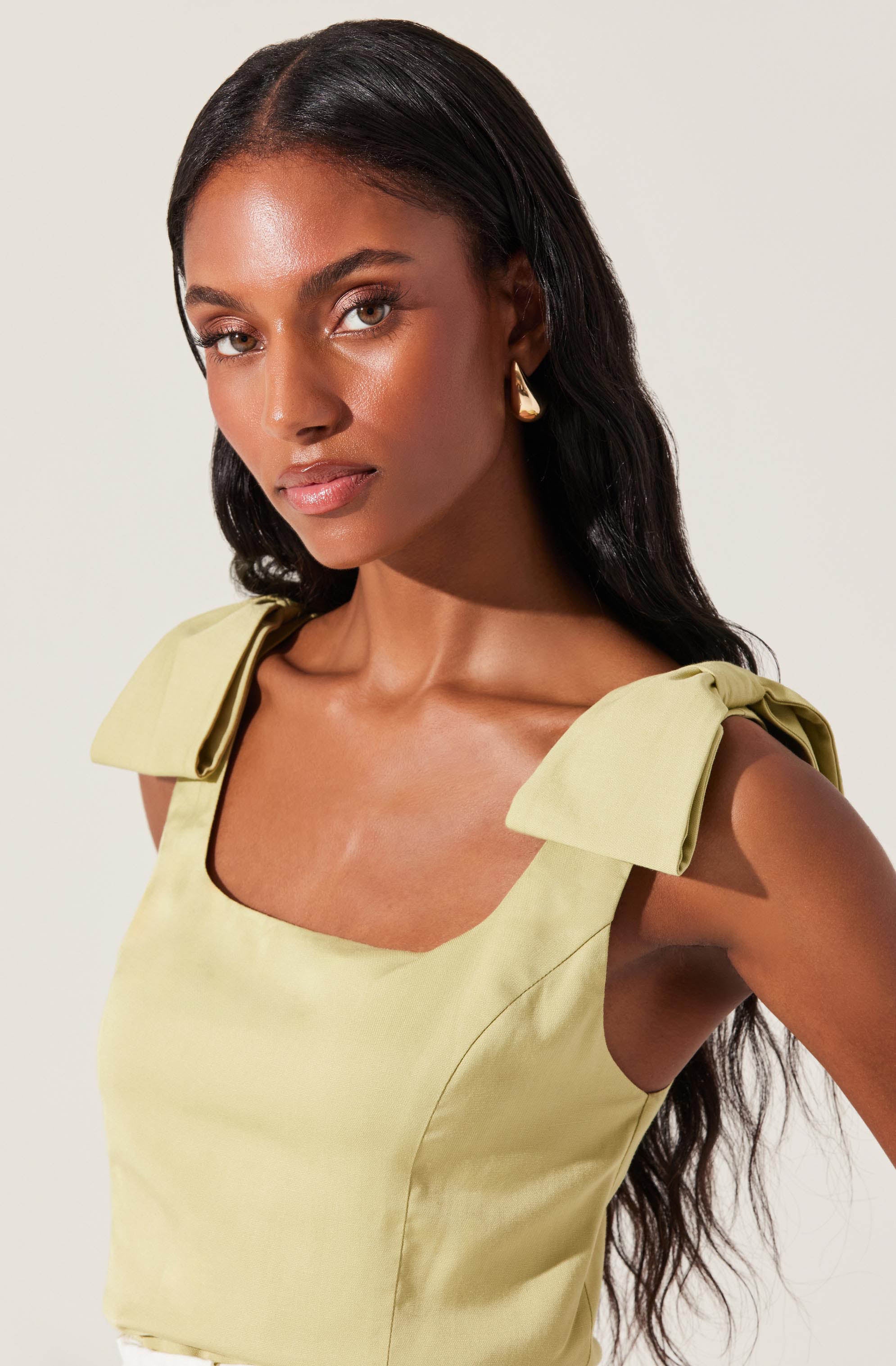 A woman is wearing a light yellow sleeveless top with shoulder bows. The top has a square neckline and is made of a smooth, shiny fabric.