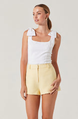 A woman is wearing a sleeveless white top with large bow details on the shoulder straps and a square neckline. She pairs the top with high-waisted, light yellow textured shorts.