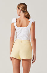 A woman is wearing a sleeveless white top with wide shoulder straps and a square neckline. She pairs it with high-waisted, light yellow textured shorts that have a back pocket and a fitted waistband.