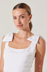A woman is wearing a sleeveless white top with large bow details on the shoulders. The top has a rounded neckline and appears to be made of a smooth, structured fabric.