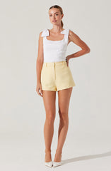 A woman is wearing a white sleeveless top with shoulder bow details and a square neckline, paired with light yellow textured shorts with a high waist and front zipper. She is also wearing white pointed-toe shoes.