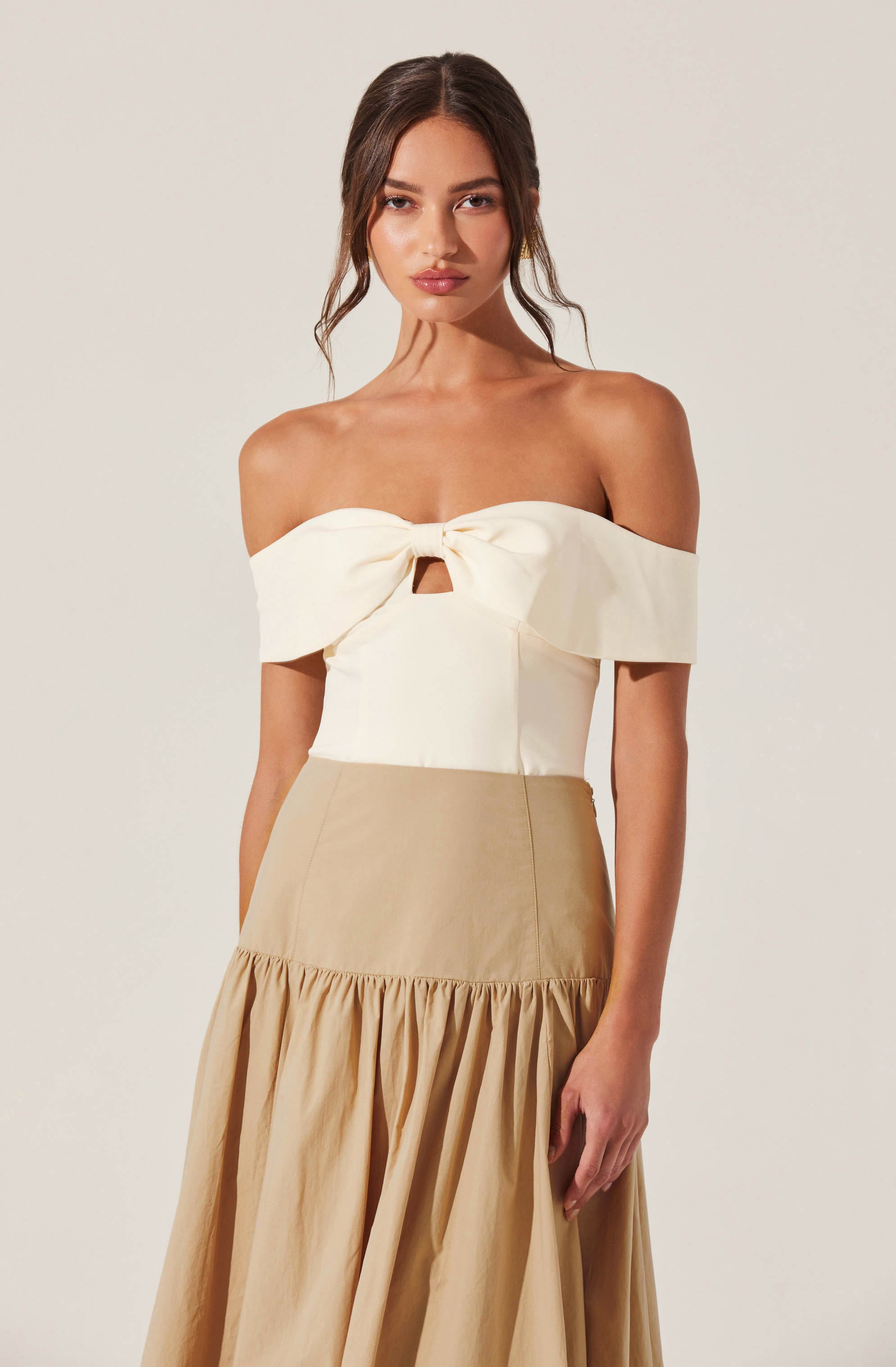 A woman is wearing an off-the-shoulder cream-colored top with a large bow and a small keyhole cutout in the center. The top has a structured fit and wide, folded-over sleeves. She is also wearing a high-waisted, beige skirt with a gathered, ruffled hem that extends to the floor.