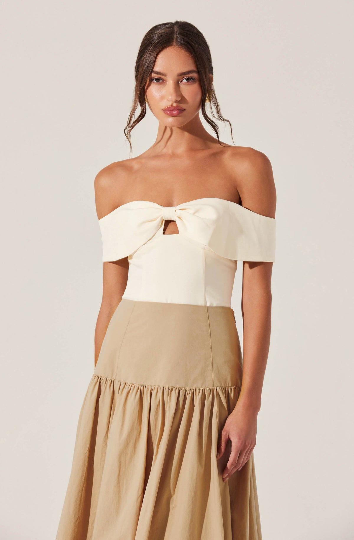 A woman is wearing an off-the-shoulder cream-colored top with a large bow and a small keyhole cutout in the center. The top has a structured fit and wide, folded-over sleeves. She is also wearing a high-waisted, beige skirt with a gathered, ruffled hem that extends to the floor.