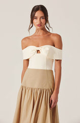 A woman is wearing an off-the-shoulder cream-colored top with a large bow and a small keyhole cutout in the center. The top has a structured fit and wide, folded-over sleeves. She is also wearing a high-waisted, beige skirt with a gathered, ruffled hem that extends to the floor.