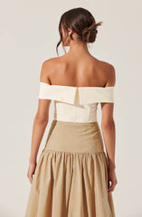 A woman is wearing an off-the-shoulder, white top with a wide folded collar that extends around her shoulders. The top has a zipper at the back. She pairs it with a high-waisted beige skirt that features a fitted upper section and a gathered, billowy lower section.