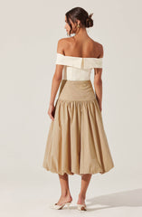 A woman is wearing an off-the-shoulder white top with a wide fold-over neckline and a back zipper. She pairs it with a high-waisted, beige, full-circle skirt that has gathered fabric at the waist creating a voluminous, balloon-like effect. She completes the outfit with white high-heeled shoes. Her hair is styled in a low, textured bun, and she is wearing earrings.