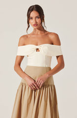 The woman is wearing an off-the-shoulder top with a bow detail at the front, made from a light, creamy fabric. She pairs it with a high-waisted beige skirt that has a gathered, voluminous design at the hem.
