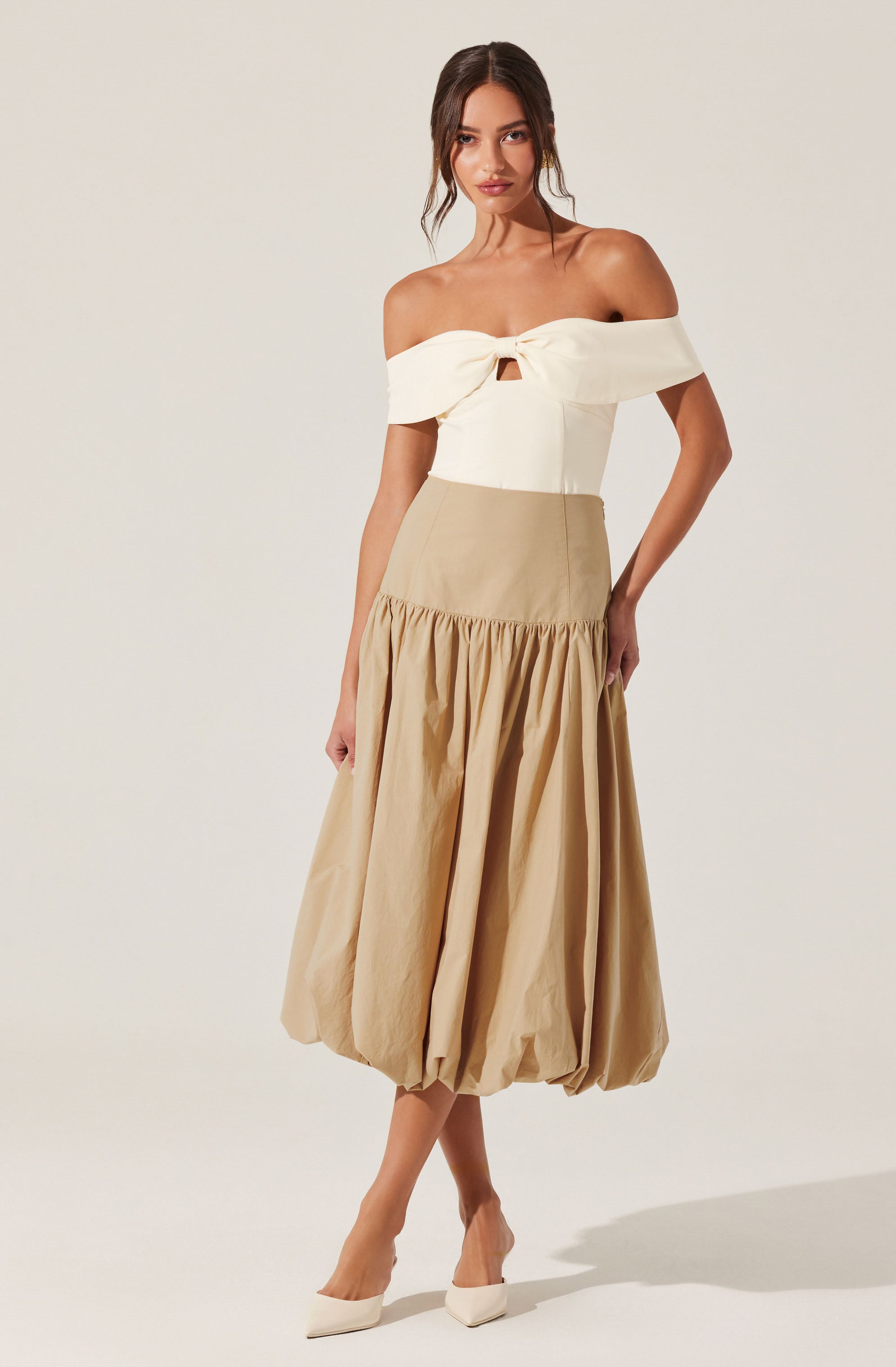 A woman is wearing an off-the-shoulder top in a light cream color with a knot detail at the center of the neckline. She pairs it with a high-waisted, beige midi skirt that has a gathered, balloon-style hem. She completes her outfit with pointed-toe white shoes.