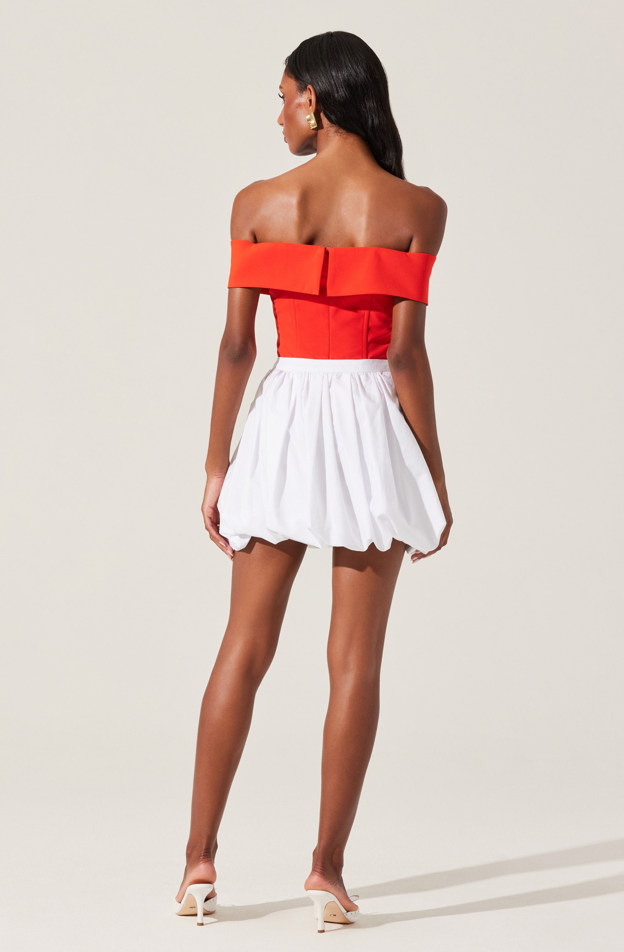 A woman is wearing an off-the-shoulder red top with a wide folded neckline and a fitted bodice, paired with a high-waisted white puffed skirt that has a gathered, balloon-like shape. She is also wearing white high-heeled sandals.
