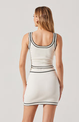 A woman is wearing a sleeveless, fitted white dress with black embroidered trim along the neckline, armholes, waist, and hem. The dress has a scoop neckline and a short length, ending above the knees.