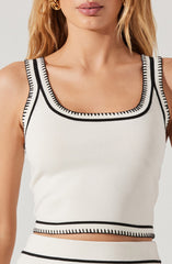 A woman is wearing a sleeveless, white crop top with black trim detailing along the neckline, armholes, and hem. The top features a rounded neckline and a fitted style.
