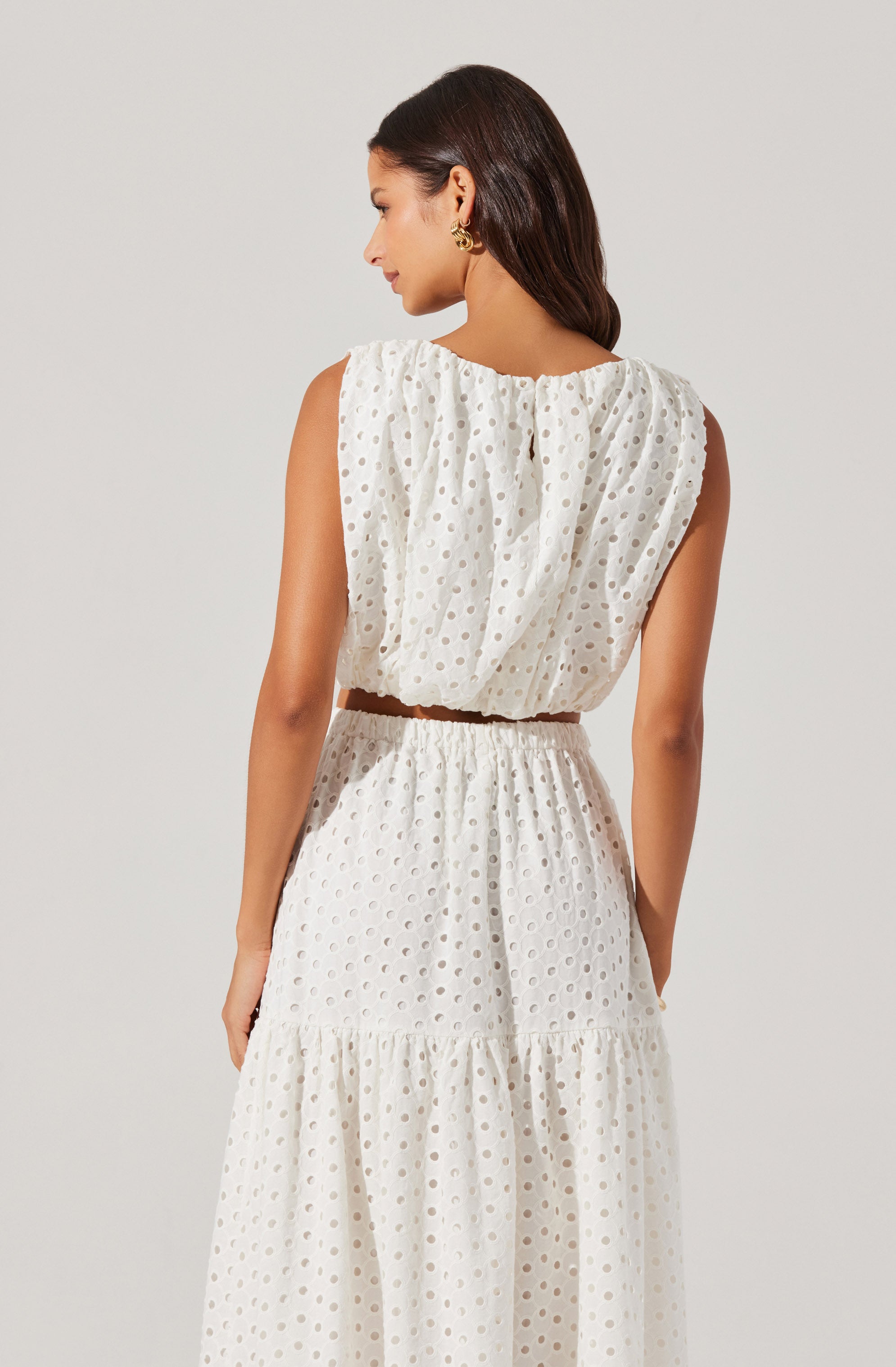 A woman is wearing a sleeveless white dress with a textured, eyelet pattern featuring circular cutouts. The dress has a relaxed fit, with gathered fabric on the top and an elastic waistband. The skirt is tiered, with the lower tier slightly longer, creating a flowing silhouette.