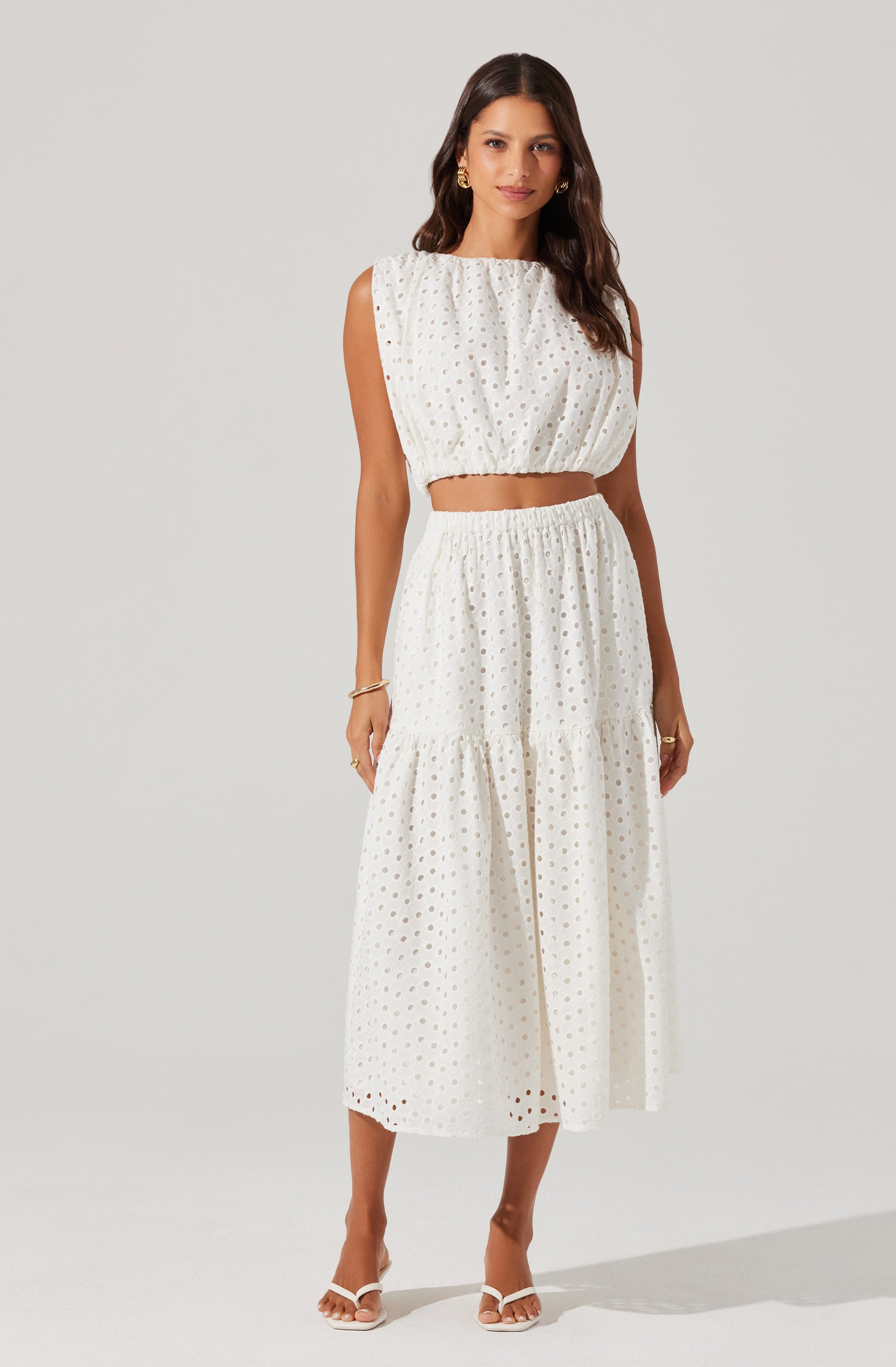 A woman is wearing a two-piece outfit made of white fabric with a cut-out eyelet pattern. The top is sleeveless with a gathered neckline and a loose, cropped fit that exposes her midriff. The matching skirt is high-waisted with an elastic waistband, a tiered design, and a midi length, flowing down to her ankles. She is also wearing white open-toe sandals.