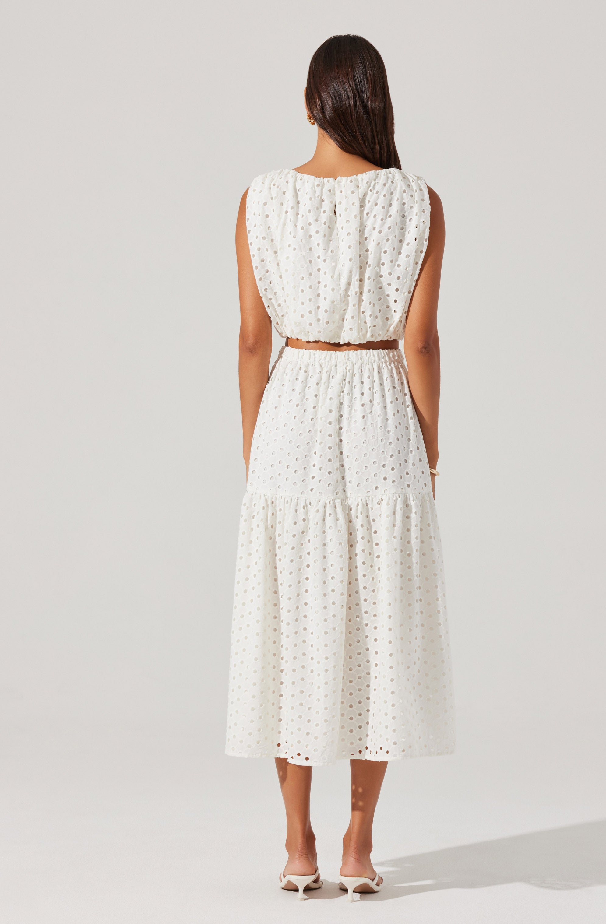 A woman is wearing a sleeveless white dress with an eyelet pattern. The dress has a loose, gathered bodice with a round neckline and a cropped waistline. The skirt is tiered, with a gathered seam at the mid-length and a flowing, ankle-length hem. She is also wearing white heeled shoes.
