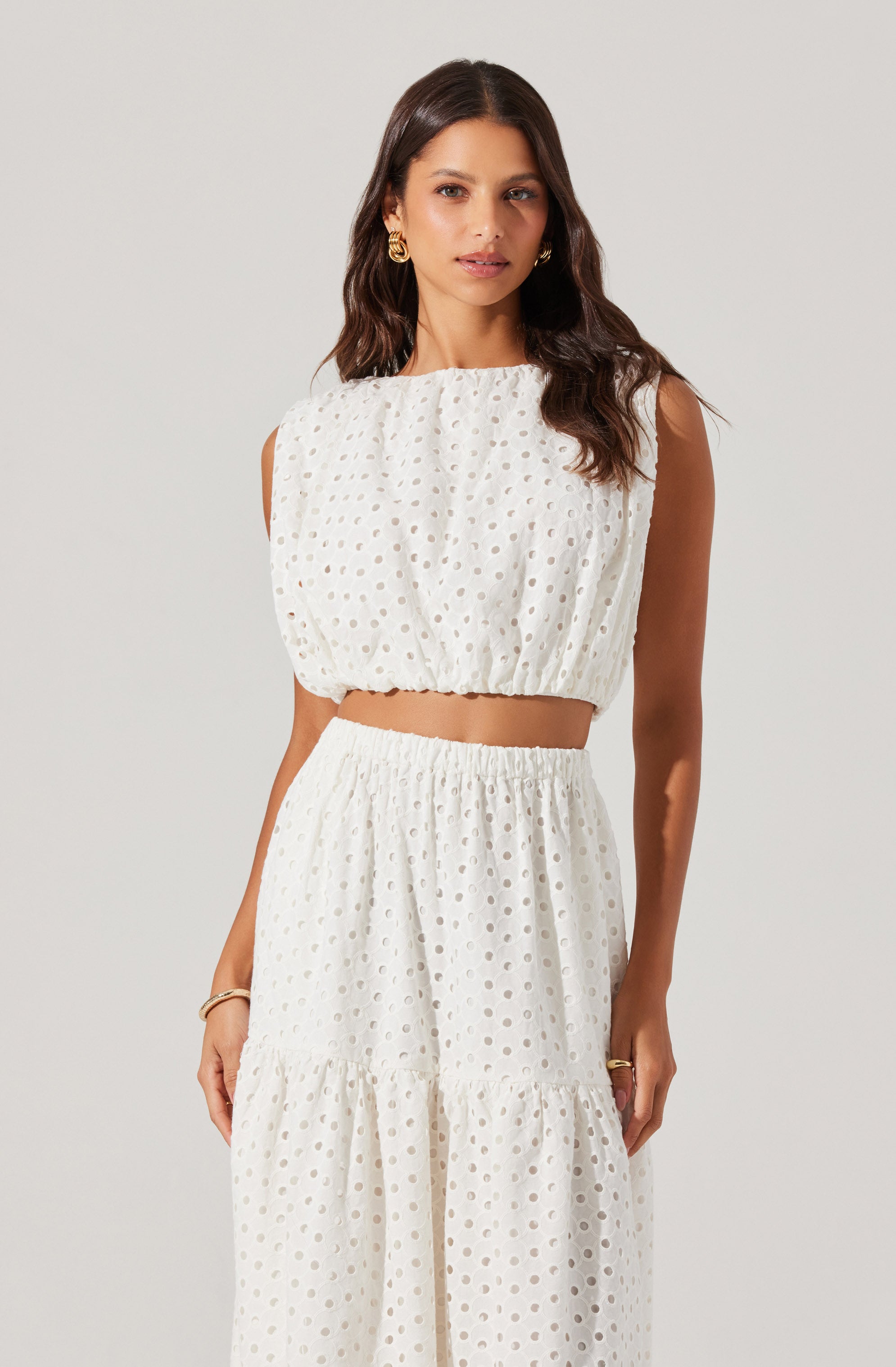 A woman is wearing a white two-piece outfit made of a lightweight, textured fabric with a polka dot pattern. The top is sleeveless, loose-fitting, and cropped, with a gathered neckline and elastic hem. The matching skirt is high-waisted, long, and flowy, with an elastic waistband and a tiered, ruffled design. She has long, wavy dark hair and is accessorized with large gold earrings, a gold bracelet, and rings.