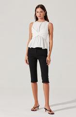 A woman is wearing a sleeveless white top with a draped, layered design and a ruffled hem, paired with black cropped pants that have small side slits at the hem. She is also wearing black open-toe high-heeled sandals and gold earrings.