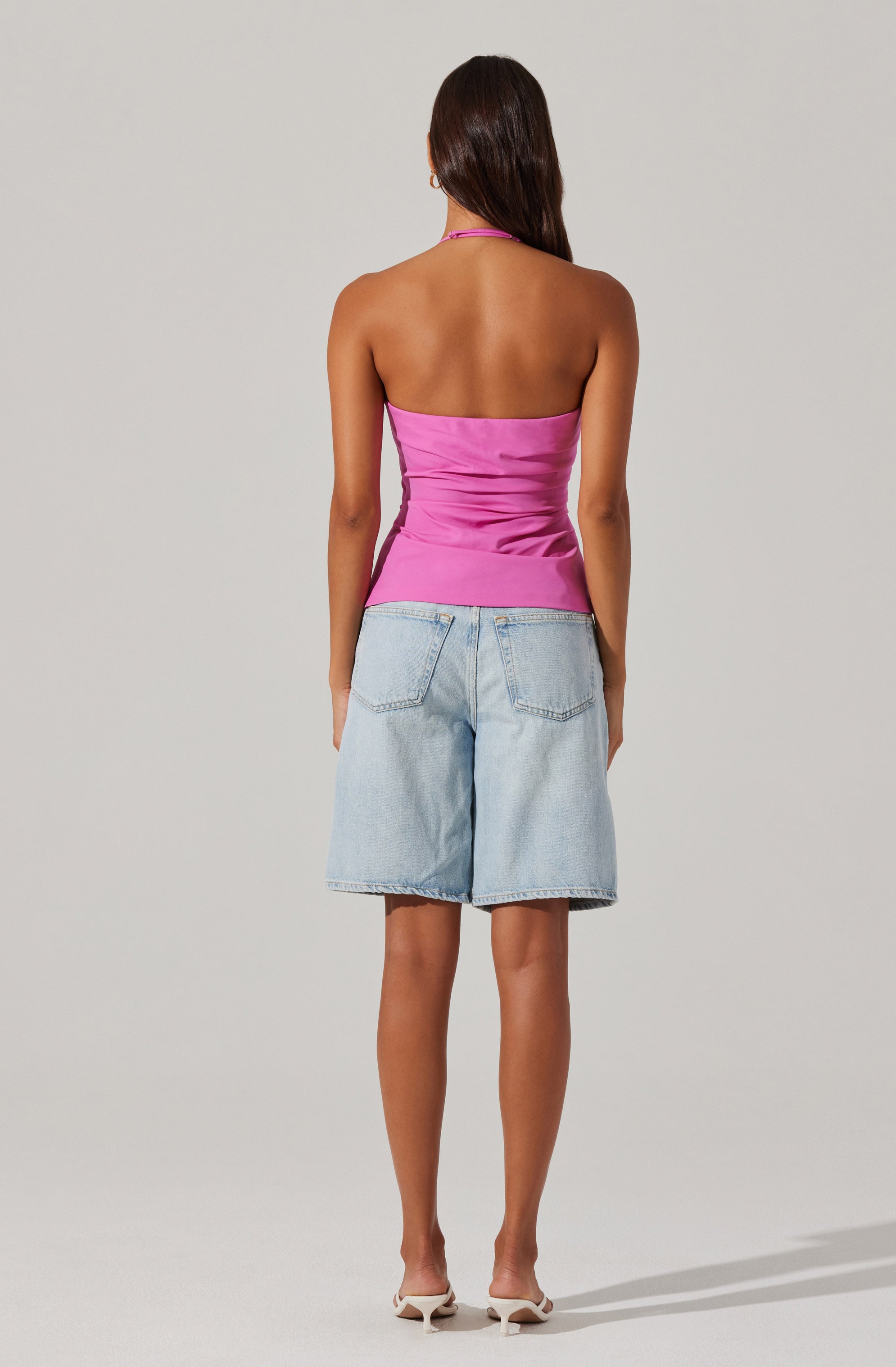 A woman is wearing a strapless, shiny pink top that is fitted and ruched around the waist. She is also wearing light-wash, knee-length denim shorts with large back pockets. She has on white high-heeled shoes.