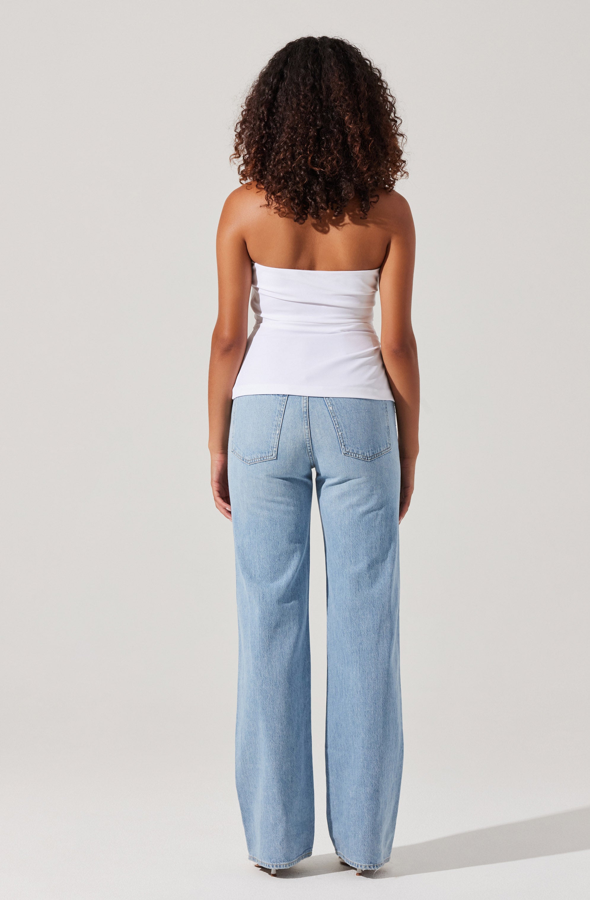 A woman is wearing a white strapless top that is fitted around the waist and flowy, light blue, high-waisted, wide-leg jeans.