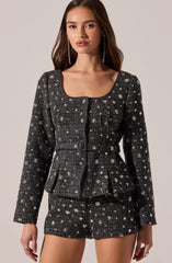 A woman is wearing a matching two-piece outfit consisting of a long-sleeved, fitted jacket and a pair of shorts, both made from a dark, textured fabric adorned with a pattern of small, light-colored floral or star-like motifs. The jacket has a round neckline, button closures down the front, and a belt at the waist. The shorts are tailored with a clean, simple design.