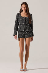 A woman is wearing a matching set consisting of a black, textured fabric blazer and shorts, both decorated with a pattern of small white stars. The blazer has a round neckline, long sleeves, and buttons down the front. The shorts are fitted and high-waisted. She is also wearing open-toed heels with a snakeskin pattern.