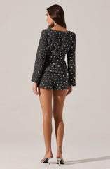 A woman is wearing a fitted blazer and matching shorts set made of dark fabric decorated with a pattern of white stars. She is also wearing high-heeled shoes with a metallic look and sparkly earrings.