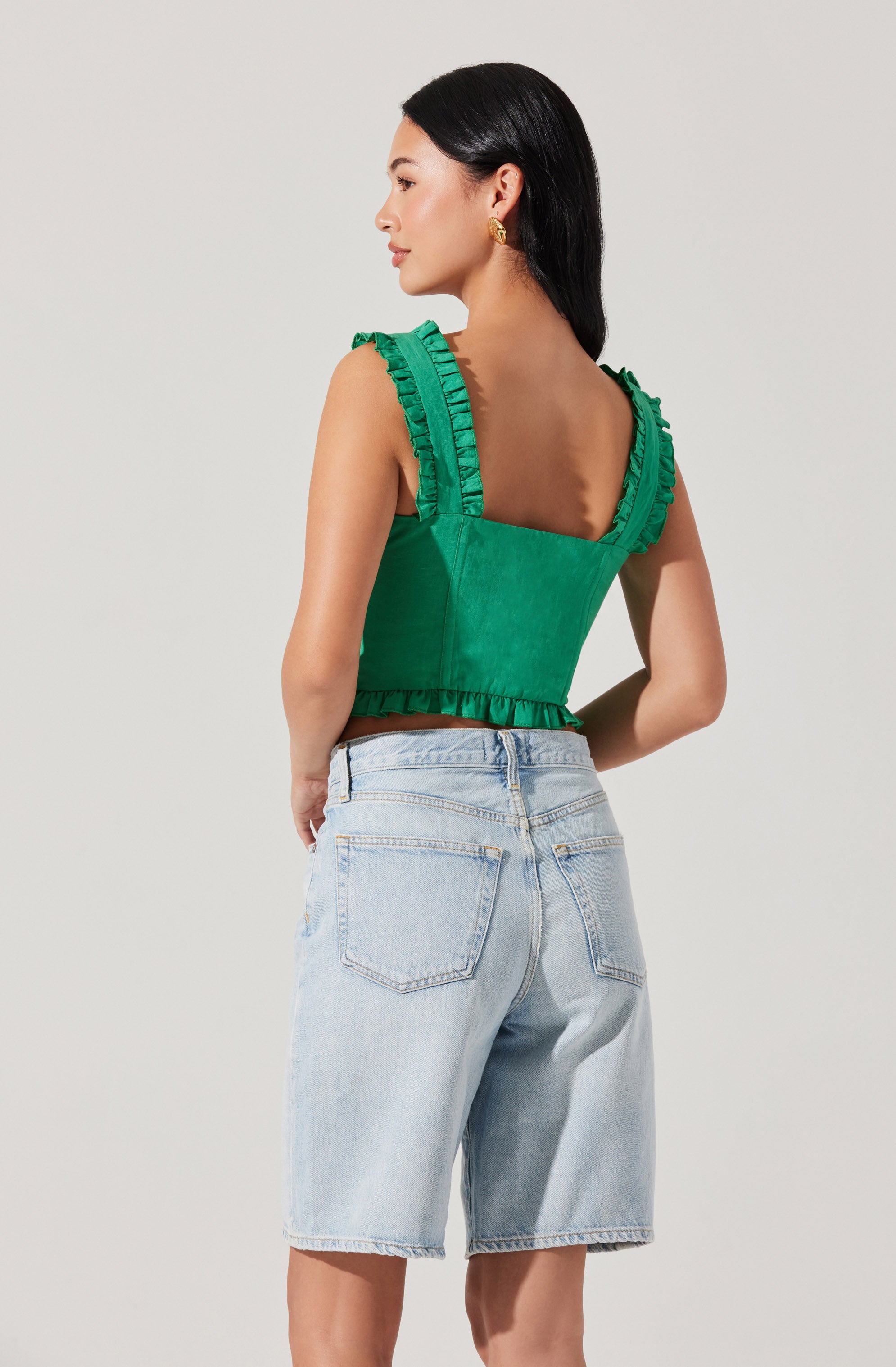 A woman is wearing a sleeveless green top with ruffled straps and a ruffled hem, paired with light-wash denim shorts.