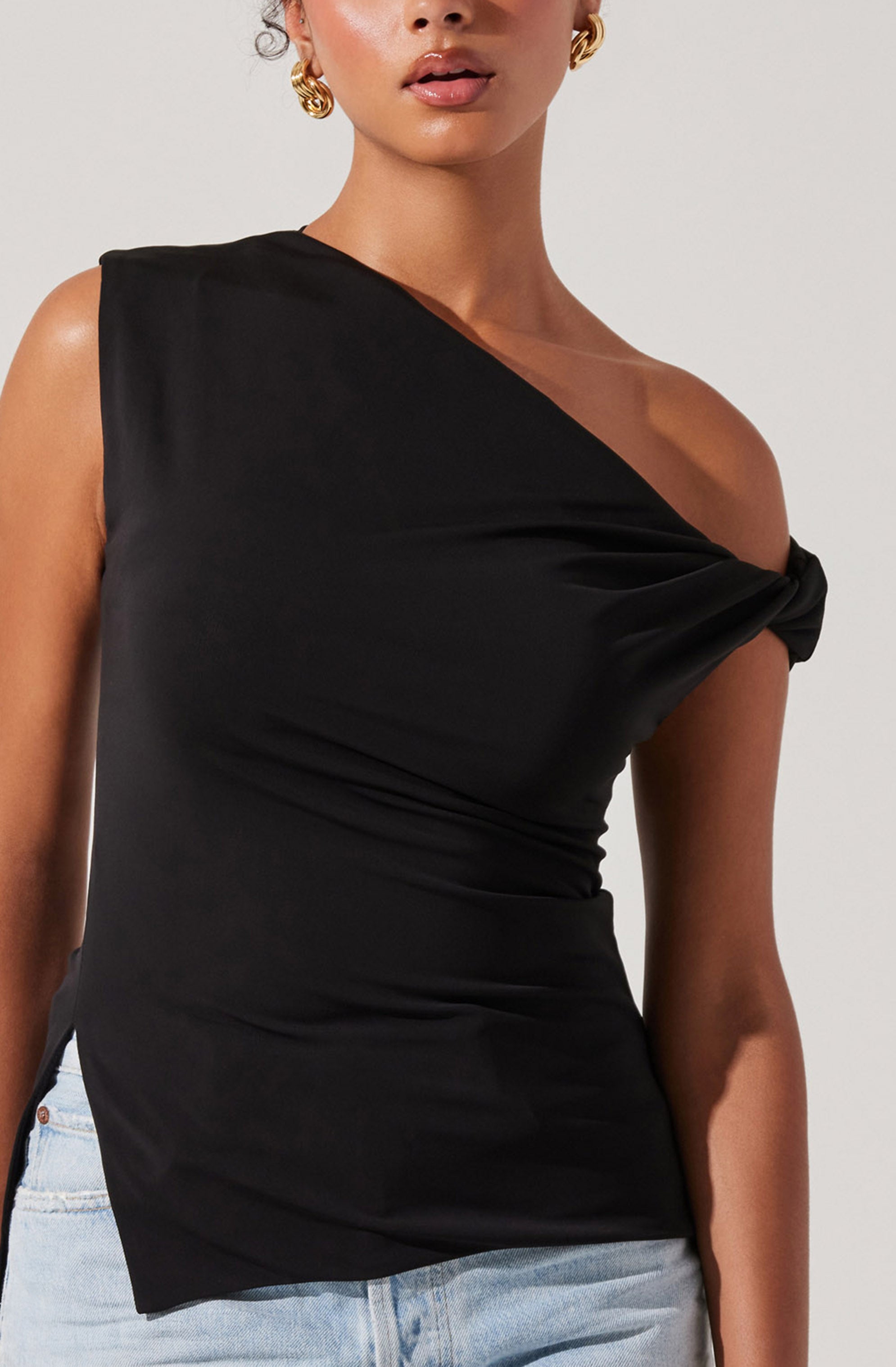 A woman is wearing a black, sleeveless top with an asymmetrical neckline. One shoulder features a knot detail, creating a draped effect across the chest. The fabric appears smooth and slightly shiny. She pairs the top with light blue denim jeans.