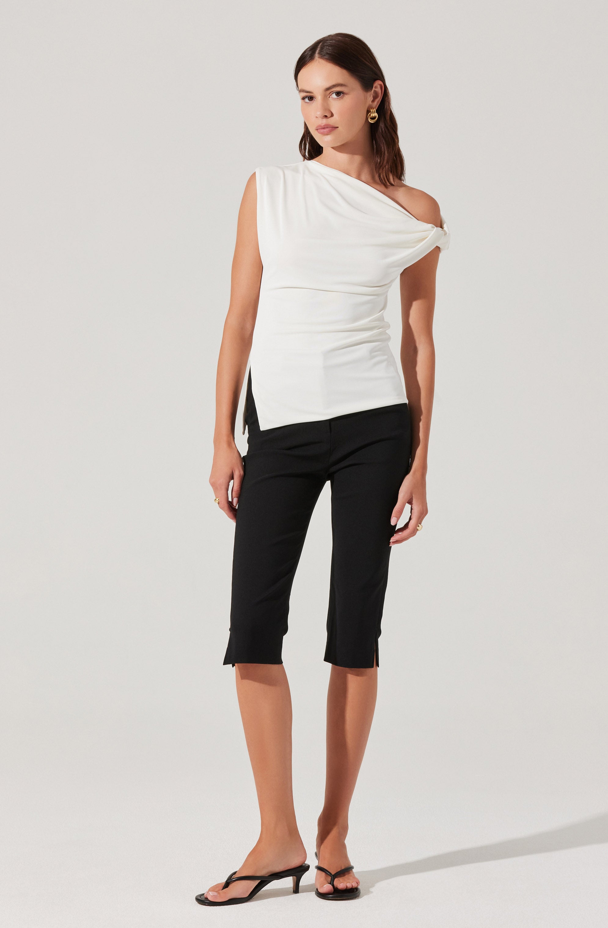 A woman is wearing a white, asymmetrical, one-shoulder top with a draped, twisted detail on the shoulder and a slit on the side. She pairs it with black, knee-length, fitted pants that have small slits at the hems. She is also wearing black open-toe high-heeled sandals and gold earrings.