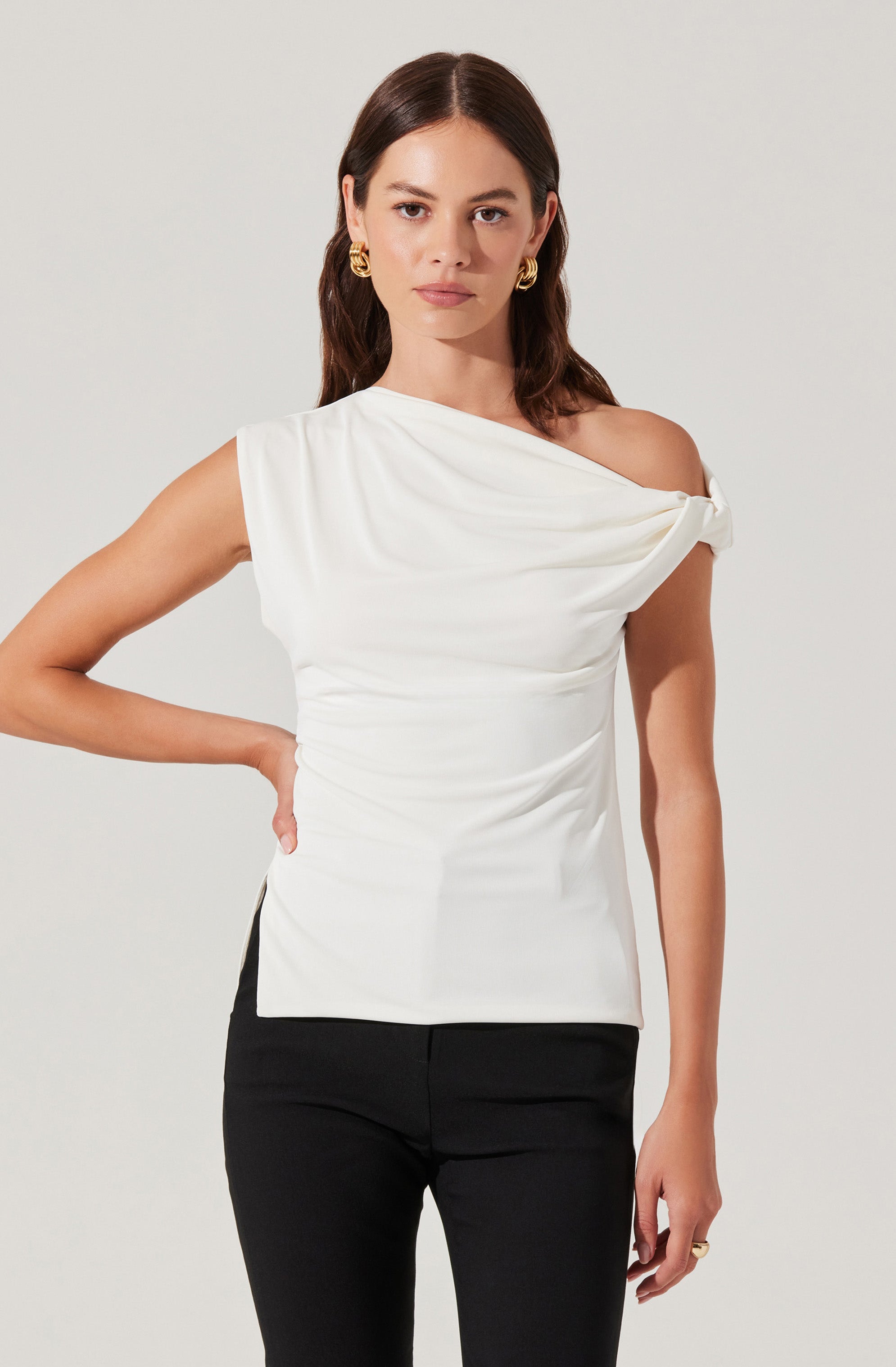 A woman is wearing a sleeveless white top with an asymmetrical design, featuring a draped, off-the-shoulder style on her right side and a twist detail at the shoulder. She pairs it with black pants and accessorizes with large gold hoop earrings and a ring on her left hand.