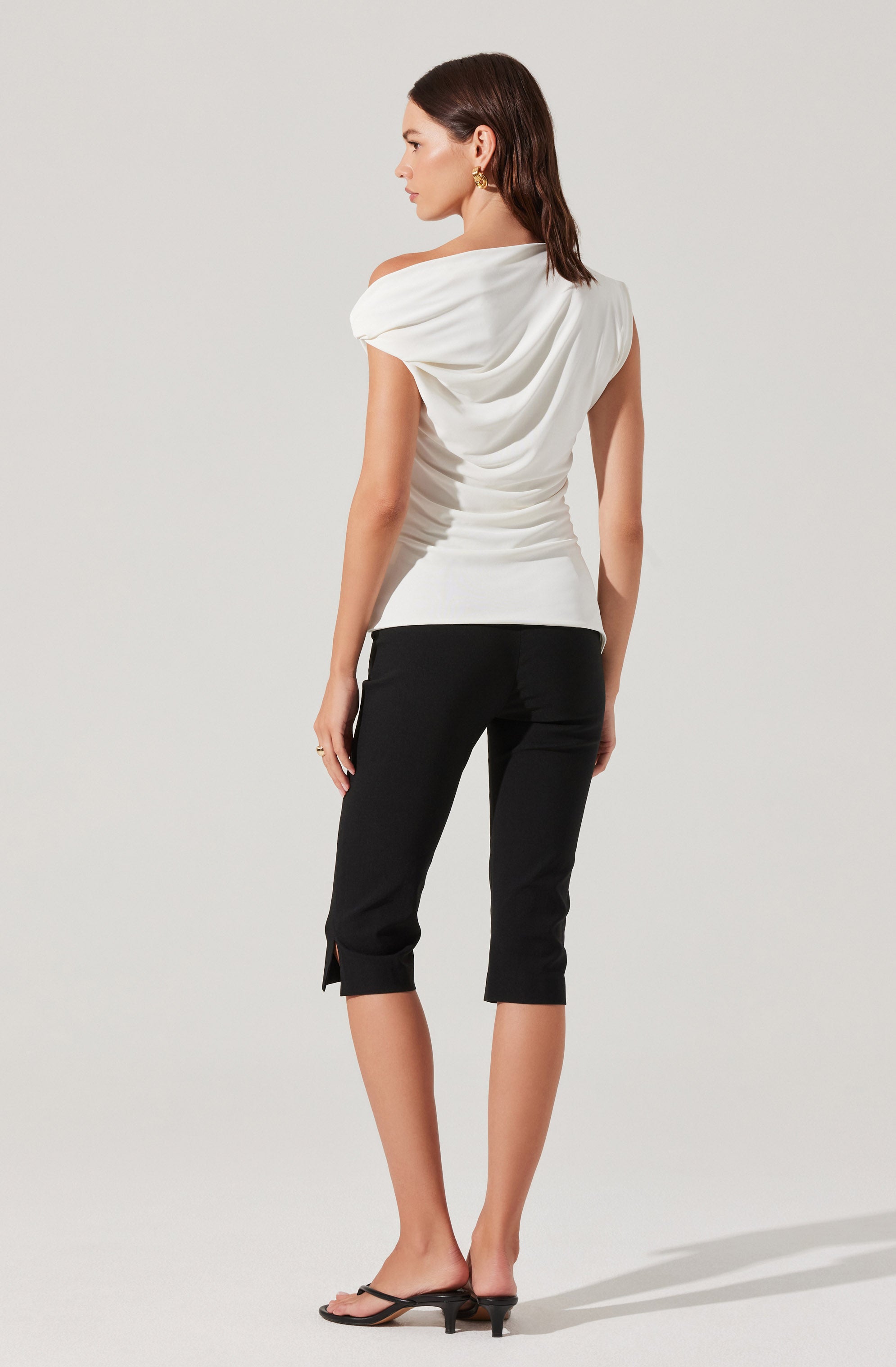 A woman is wearing a white, sleeveless top with a draped, cowl neckline and gathered fabric at the waist. She pairs it with black, cropped capri pants that have a slit at the hem on one side. She completes the outfit with black, open-toe sandals with a small heel.