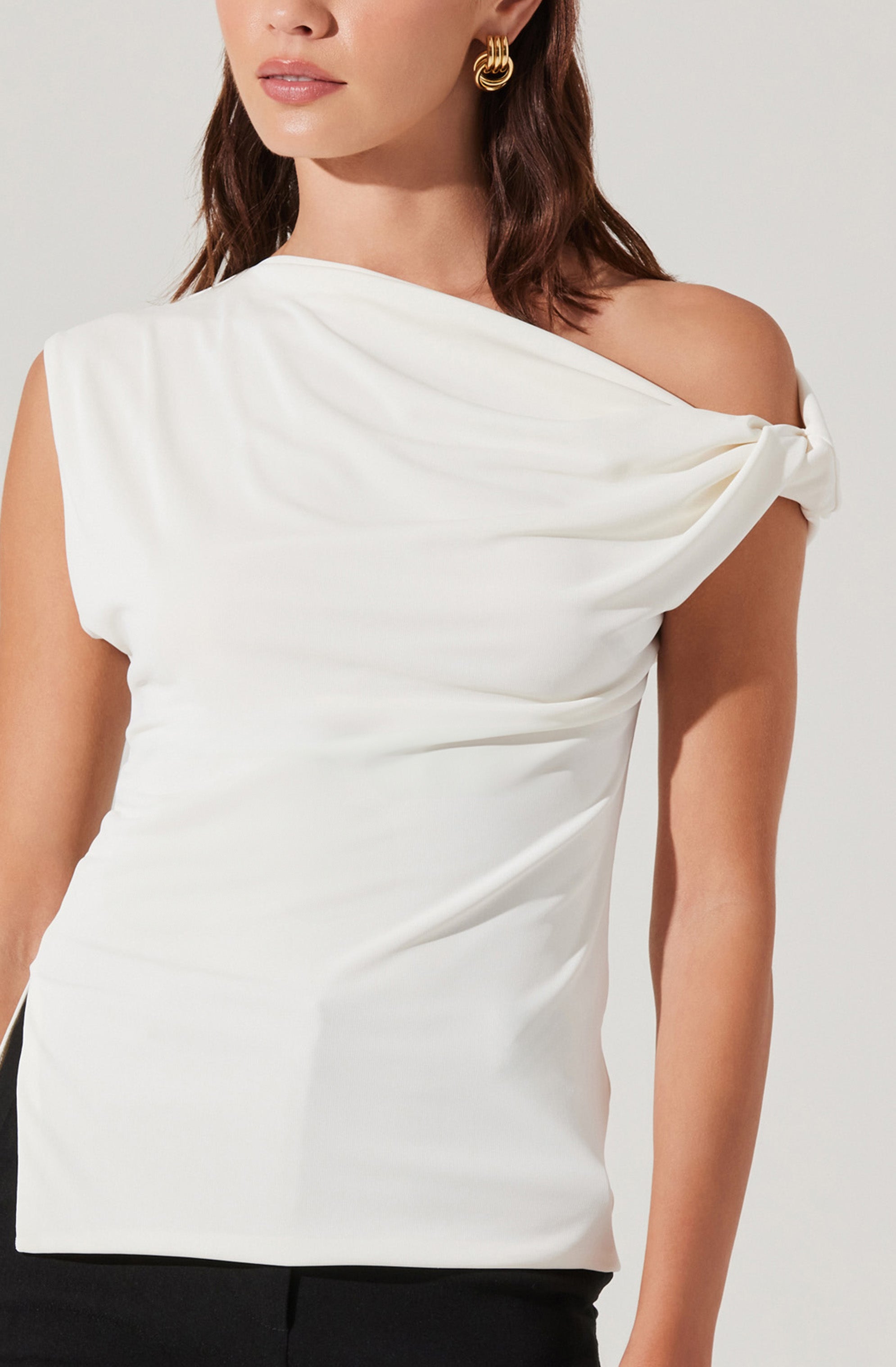 A woman is wearing a sleeveless, white asymmetrical top with a draped shoulder detail that features a knot on one side. The fabric appears smooth and slightly shiny, creating a sleek and sophisticated look. She is also wearing gold hoop earrings.