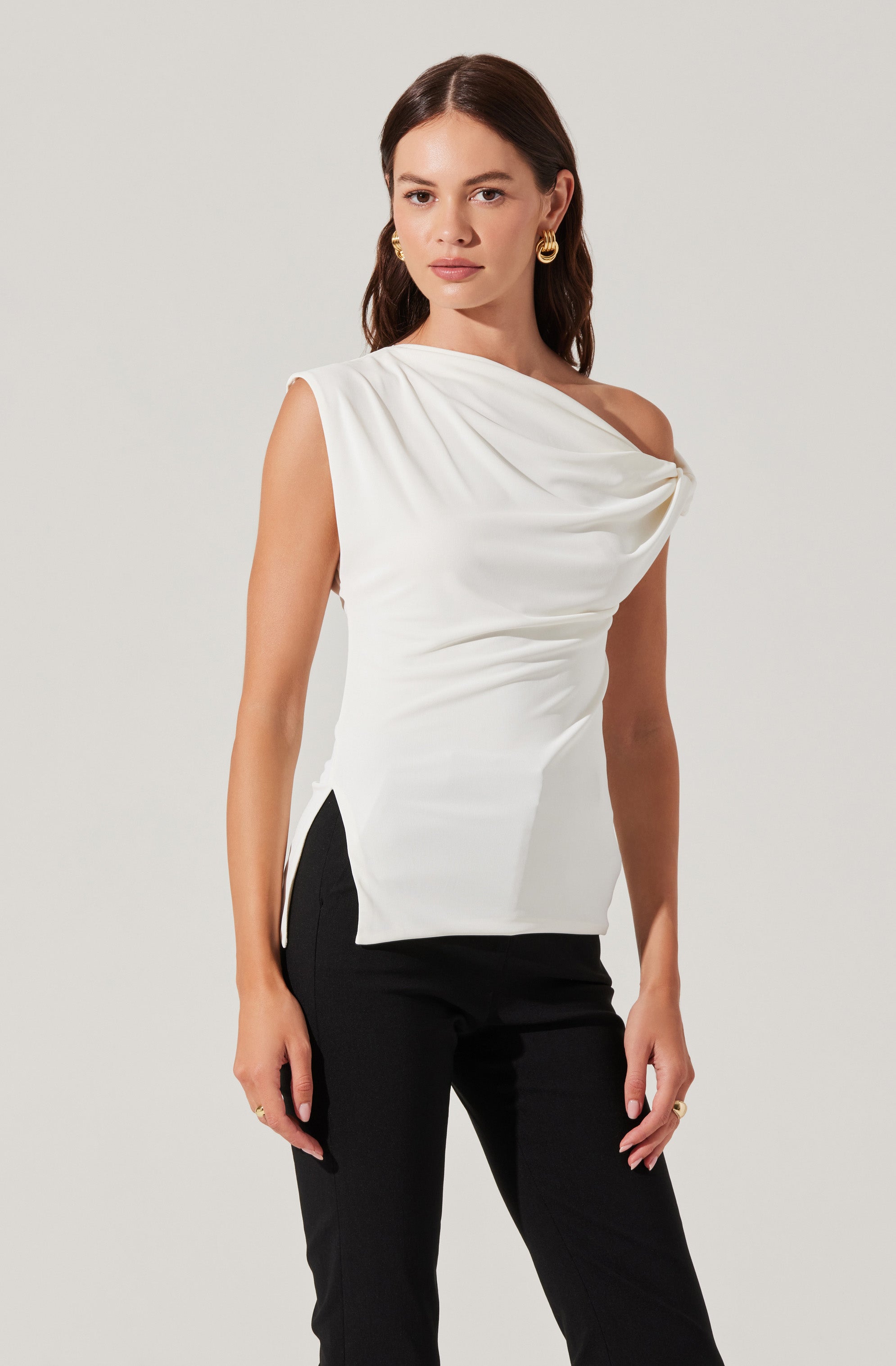 A woman is wearing a sleeveless, one-shoulder white top with draped fabric detail at the neckline. The top has a slightly asymmetrical design, with gathers creating a soft, elegant look. She is also wearing black pants with a side slit detail. She has long brown hair, gold hoop earrings, and a few rings on her fingers.