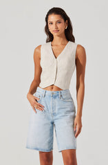 A woman is wearing a sleeveless, beige crop top with a V neckline and front buttons. She has paired it with high-waisted, light blue denim shorts that reach just above the knees, with a relaxed fit.