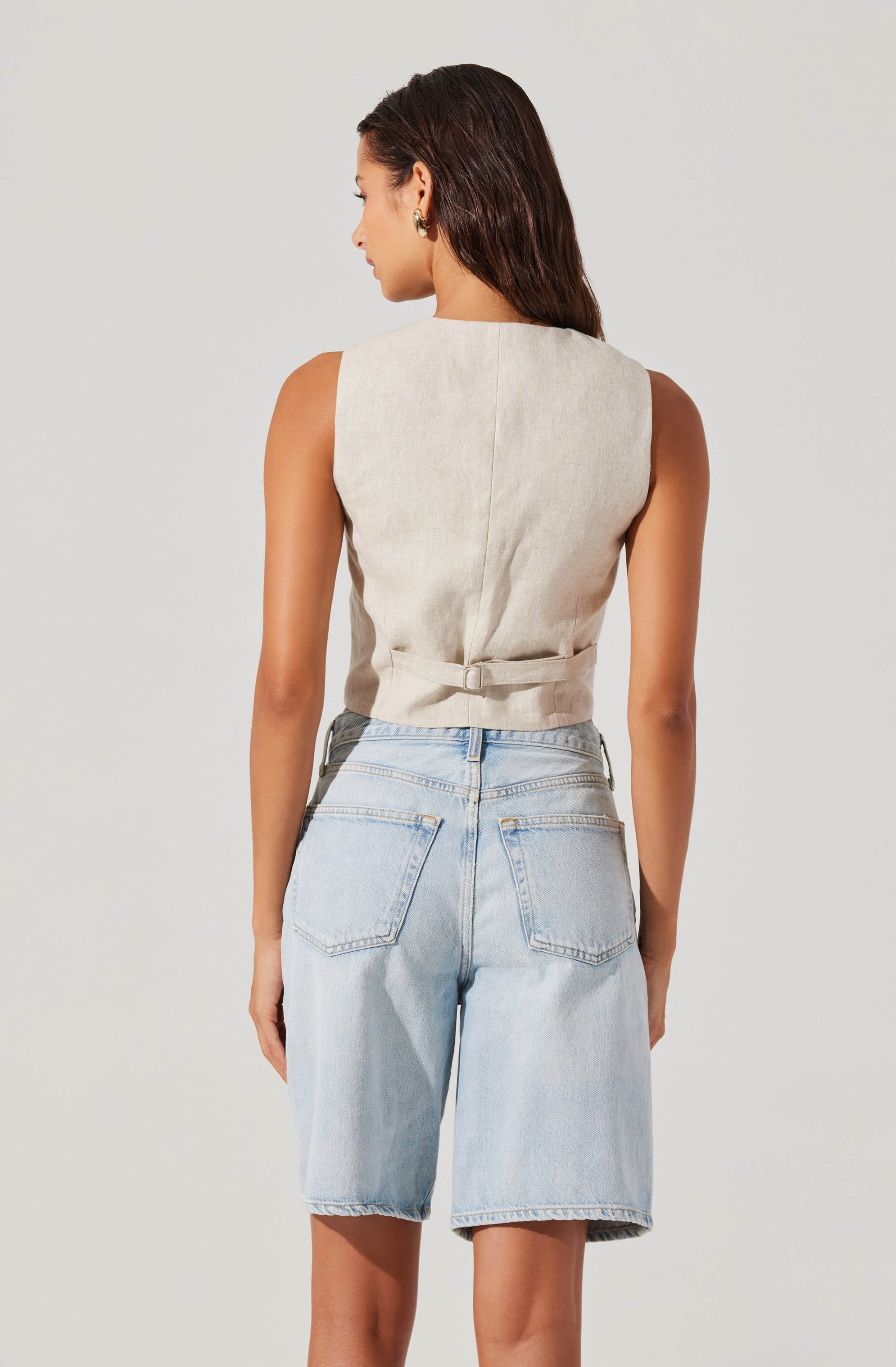 A woman is wearing a sleeveless, beige, collared vest with a waist belt and light-wash, knee-length denim shorts with back pockets.