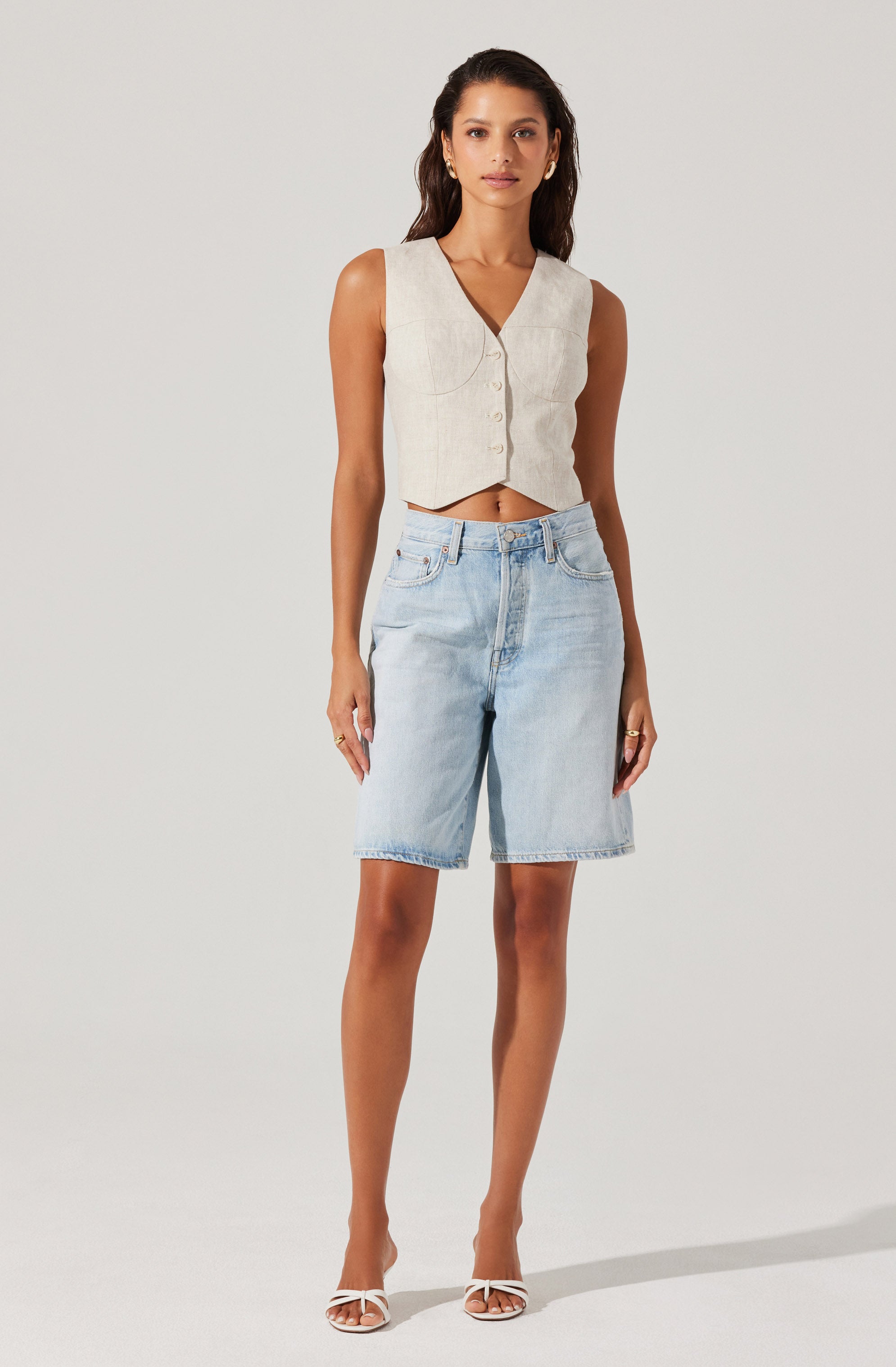 A woman is wearing a sleeveless, light beige button-up crop top with a V-neckline and a slight notch at the bottom hem. She has paired it with high-waisted, light-wash denim shorts that reach just above the knees. She is also wearing white, open-toe high-heeled sandals with a bow detail on the front.