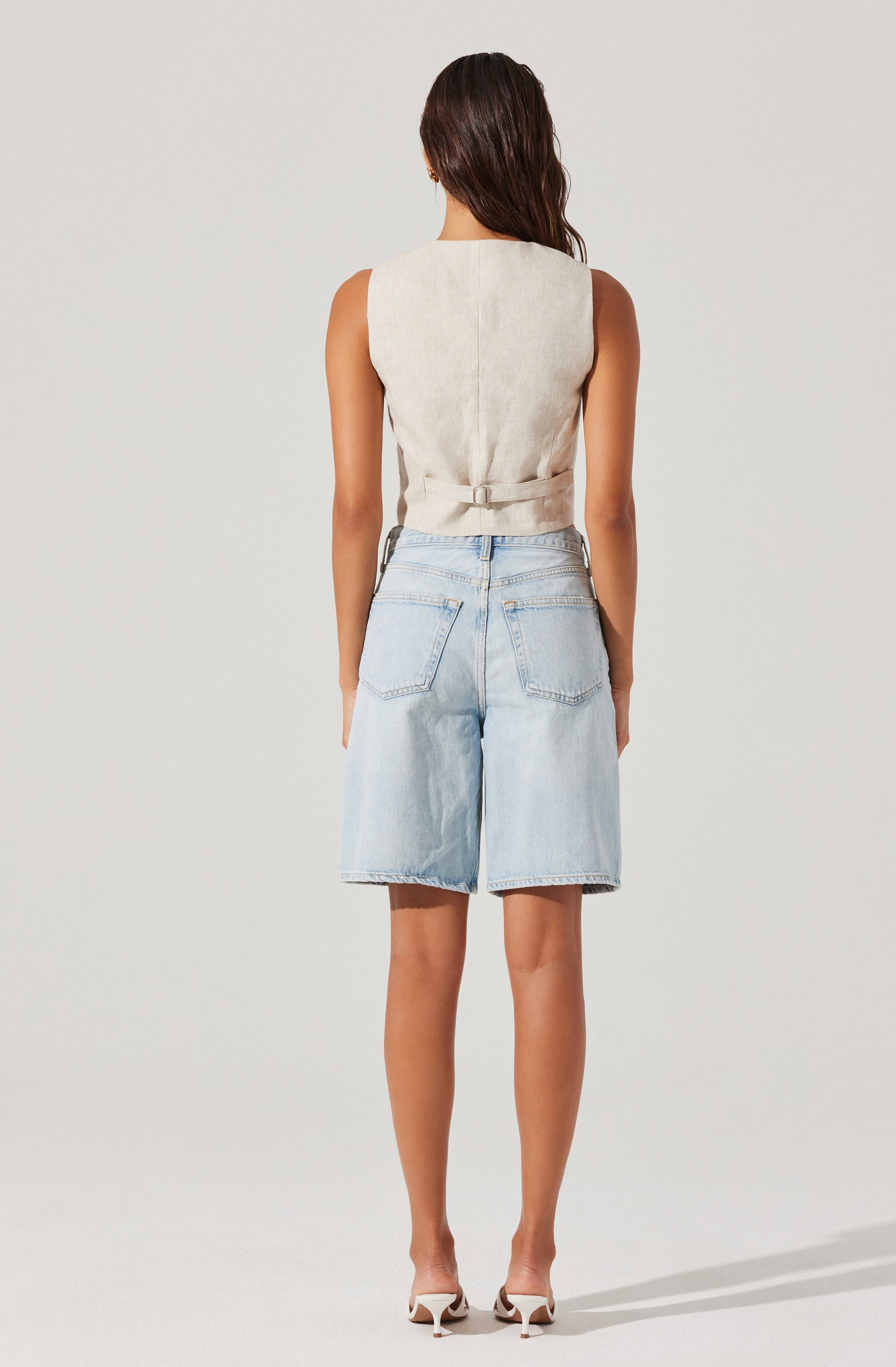 A woman is wearing a sleeveless, light beige vest with a belt at the waist, paired with high-waisted, light-wash denim shorts that have large back pockets. She is also wearing white high-heeled shoes.