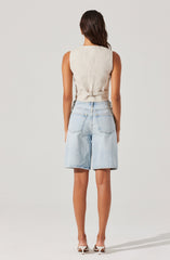 A woman is wearing a sleeveless, light beige vest with a belt at the waist, paired with high-waisted, light-wash denim shorts that have large back pockets. She is also wearing white high-heeled shoes.