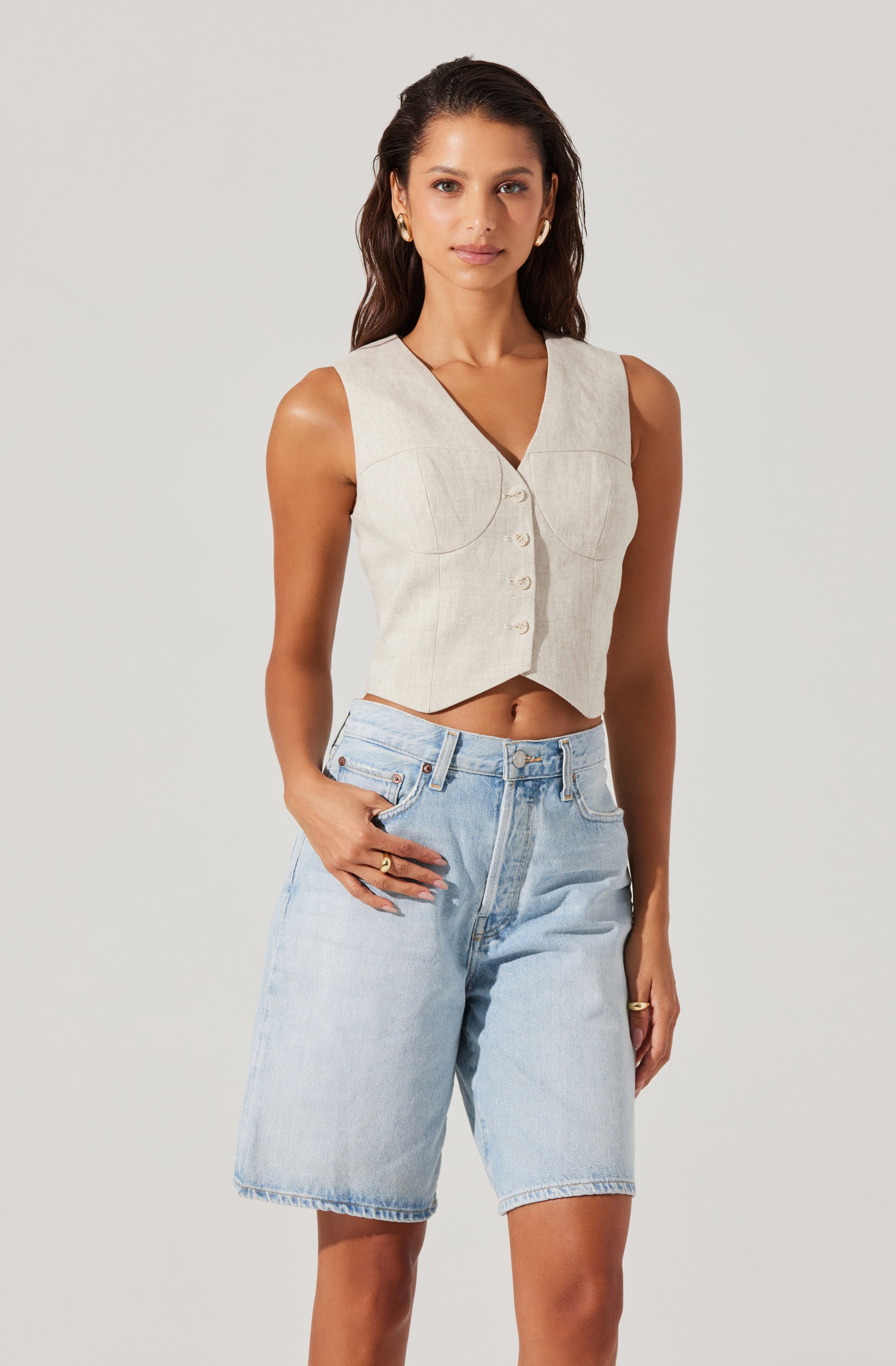 A woman is wearing a sleeveless, beige, button-up vest with a V-neckline and padded shoulders. She has paired it with light blue, high-waisted denim shorts that are loose-fitting and end just above the knees.