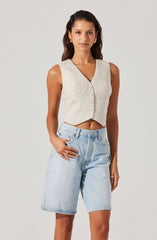 A woman is wearing a sleeveless, beige, button-up vest with a V-neckline and padded shoulders. She has paired it with light blue, high-waisted denim shorts that are loose-fitting and end just above the knees.