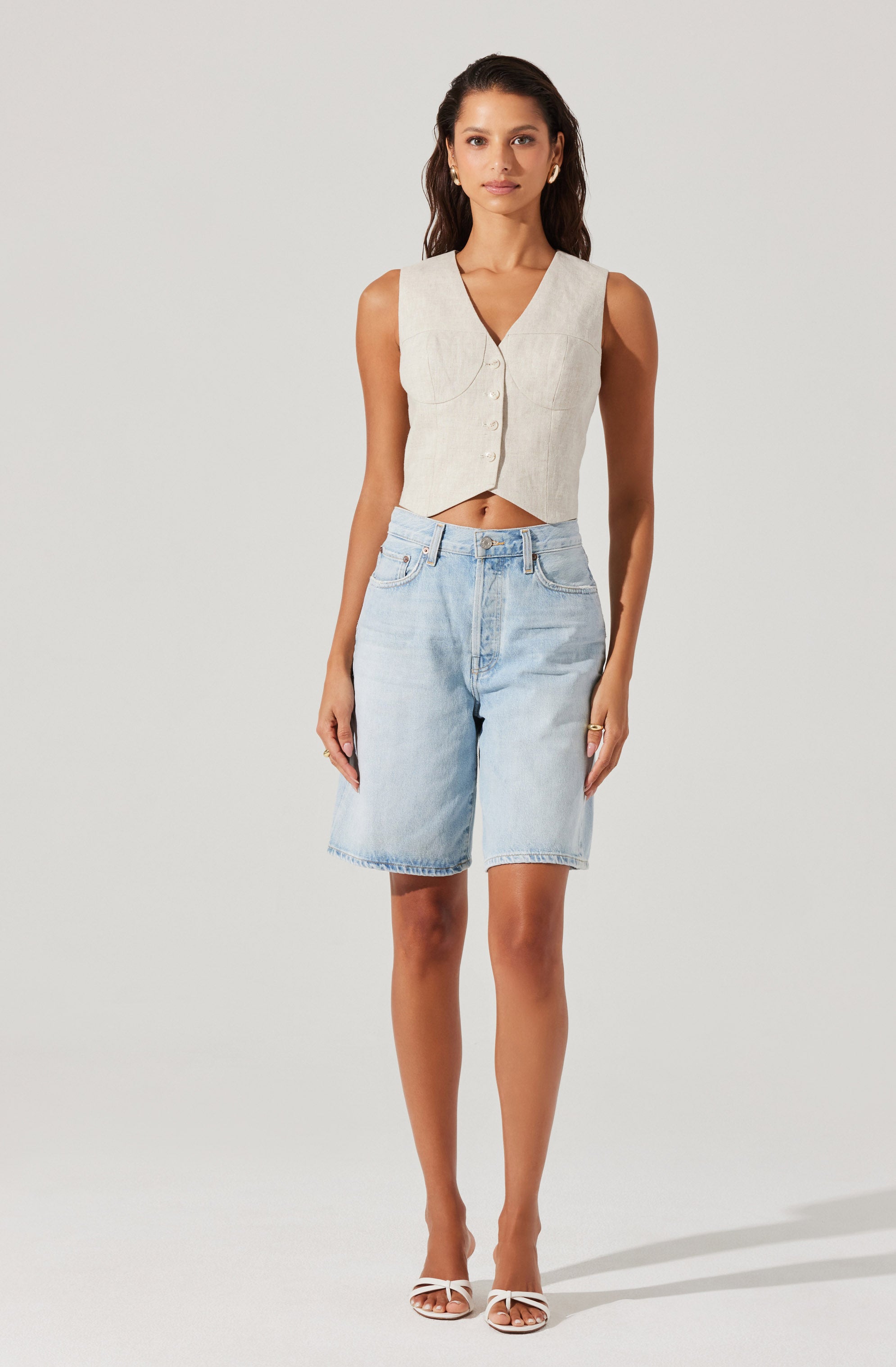 A woman is wearing a sleeveless, V-neck, button-up vest top in a light beige color. She pairs it with high-waisted, light-wash denim shorts that reach just above the knees. She completes the outfit with white strappy high heels.