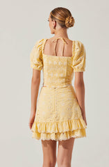 The woman is wearing a matching yellow embroidered two-piece outfit. The top has puffed sleeves and a square neckline with tie closures at the back. The skirt features a ruffled hem and eyelet embroidery throughout.