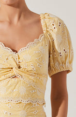 A woman is wearing a yellow dress with intricate white embroidery featuring floral and butterfly patterns. The dress has puffed short sleeves with elastic at the hem and a sweetheart neckline with ruffled lace trim. There is a ruched detail in the center of the bodice with a knot, and the fabric appears lightweight and textured.