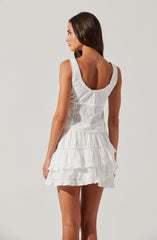 The dress is a sleeveless, white summer dress with a scoop neckline adorned with lace trim. The bodice features eyelet embroidery with floral and geometric patterns. The skirt has multiple layers of ruffled fabric, creating a tiered, textured look. The overall style is light, airy, and casual, suitable for warm weather.