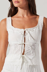 A woman is wearing a sleeveless white dress with a scoop neckline, featuring lace trim around the neckline and armholes. The dress has vertical pleats on the upper part and decorative eyelet embroidery on the lower part. It is embellished with small ribbons tied into bows along the front.